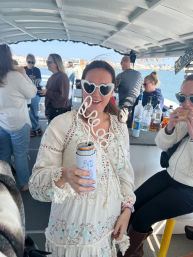 Person in heart-shaped sunglasses and a boho white embroidered dress sipping a canned drink through a curly straw aboard a sunny harbor boat party with friends and bottles at the bar.