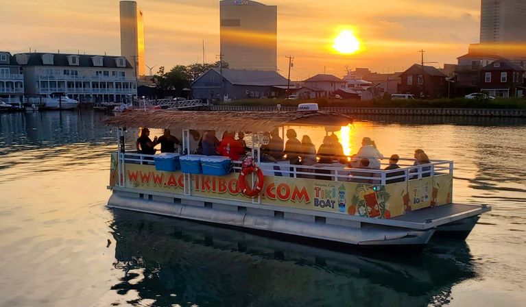 Tiki-style pontoon boat with passengers cruising a marina at golden sunset, coastal skyline and warm reflections on calm harbor water.