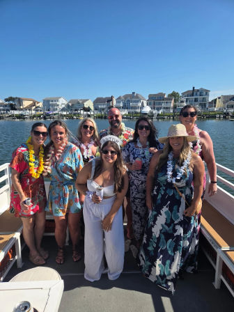 Sunny summer bachelorette on a boat — group of friends in tropical prints and leis posing near coastal waterfront homes, bride in white with a 'bride' headband holding a drink.