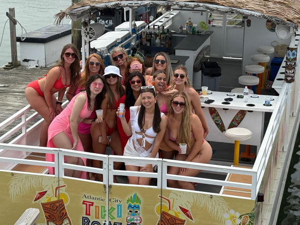 Group of smiling women in colorful swimsuits posing on a tiki-style party boat docked at an Atlantic City waterfront, with a small bar, stools and tropical decorations