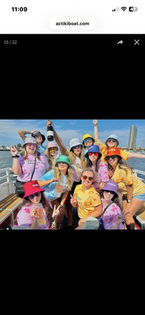 Group of friends on a sunny boat party by a marina, wearing colorful tie-dye shirts and bucket hats, holding a champagne bottle and flashing peace signs with blue sky and waterfront skyline in the background.