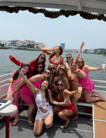 Cheerful group of women in pink swimsuits and a sash-clad bride-to-be posing with drinks on a boat in a sunny coastal bay with waterfront homes and a marina in the background.