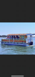 Passengers enjoying a tiki-style pontoon boat with a thatched roof and colorful signage, cruising calm blue coastal waters on a sunny day.