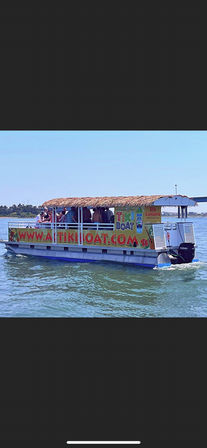 Passengers enjoying a tiki-style pontoon boat with a thatched roof and colorful signage, cruising calm blue coastal waters on a sunny day.