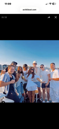 Eight friends enjoying a sunny summer boat party, holding drinks on a deck with coastal homes and clear blue sky in the background