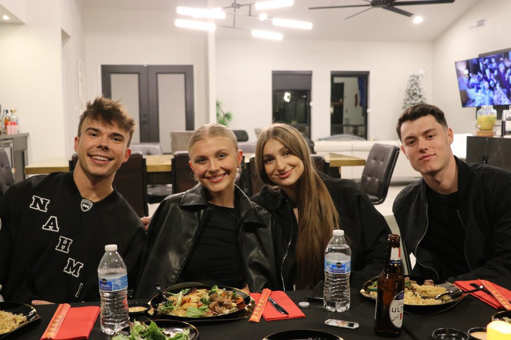Four friends smiling at a casual indoor dinner party in a modern open-plan home — plates of food, bottled water and beer on a black-tablecloth dining table, contemporary chandelier and living room in the background.