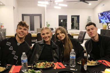 Four friends smiling at a casual indoor dinner party in a modern open-plan home — plates of food, bottled water and beer on a black-tablecloth dining table, contemporary chandelier and living room in the background.