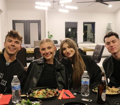 Four friends smiling at a casual indoor dinner party in a modern open-plan home — plates of food, bottled water and beer on a black-tablecloth dining table, contemporary chandelier and living room in the background.