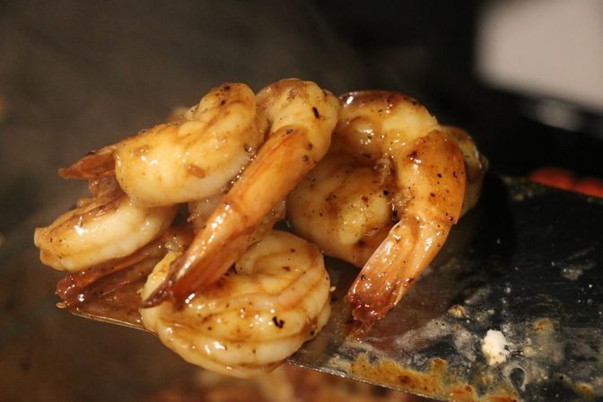 Close-up of golden-brown, seasoned sautéed shrimp glazed in a garlic-butter sauce resting on a metal spatula over a hot pan