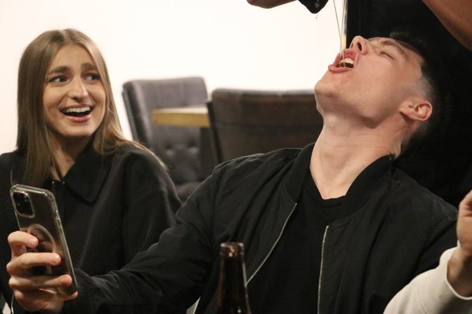 Two friends at an indoor gathering — a young man tilts his head back as a drink is poured into his mouth while a smiling woman beside him holds a smartphone and laughs.