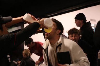 Indoor party scene with friends laughing as a man chugs beer poured from bottles into his mouth, lively celebration among young adults.