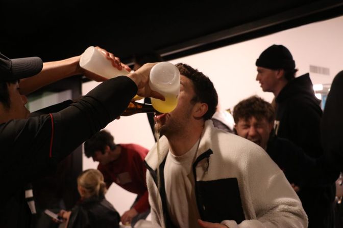 Indoor party scene with friends laughing as a man chugs beer poured from bottles into his mouth, lively celebration among young adults.