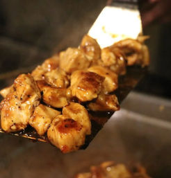 Close-up of glossy, caramelized grilled chicken bites on a metal spatula over a sizzling restaurant grill