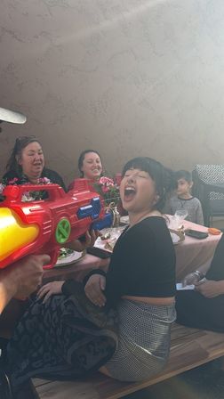 Person laughing with mouth open as someone playfully aims a large colorful water gun at an outdoor patio family gathering, guests seated at a pink-tablecloth table with flowers and plates.