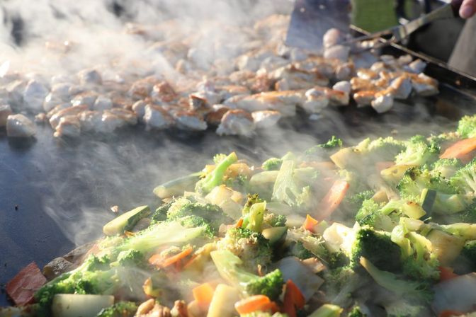 Steaming stir-fried broccoli, carrots and onions sizzling on a flat-top grill with golden grilled chicken pieces in the background at an outdoor food stall
