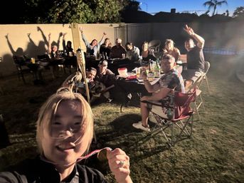 Selfie of a smiling person holding a medal at an evening suburban backyard party, friends gathered around a long table with drinks, tiki torch and pool visible in the background.