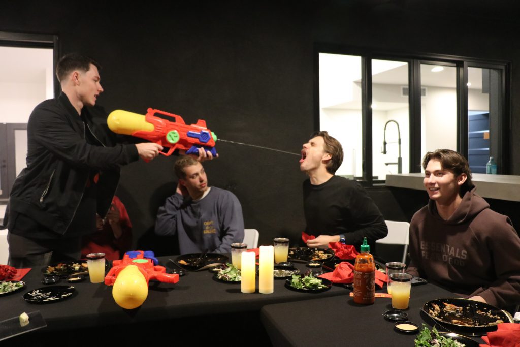 Playful indoor dinner party in a modern home: friends at a dining table with plates, candles and drinks while one person fires a large colorful water gun, spraying water into another’s open mouth.