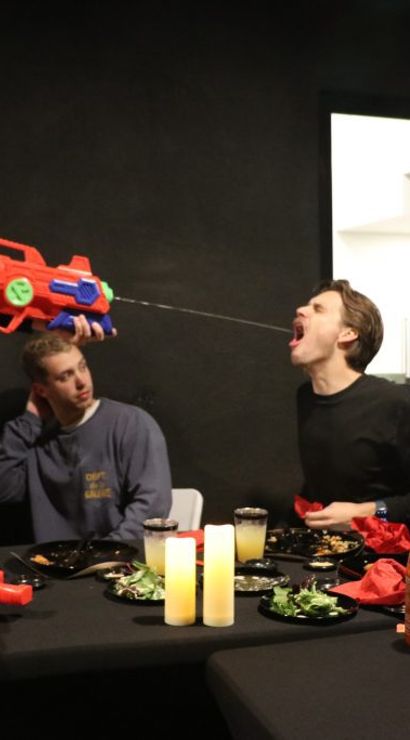 Playful indoor dinner party in a modern home: friends at a dining table with plates, candles and drinks while one person fires a large colorful water gun, spraying water into another’s open mouth.