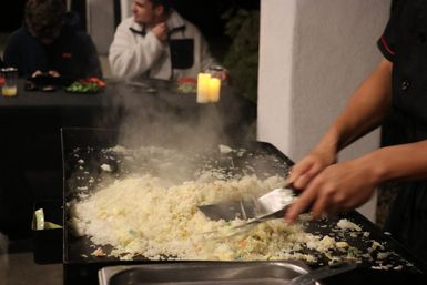 Hands using spatulas to stir steaming fried rice on a large flat-top griddle at an outdoor nighttime gathering, with blurred diners and candles in the background.