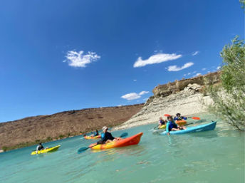 Group of colorful kayaks with paddlers on a sunlit turquoise canyon lake, paddling along sandstone cliffs beneath a clear blue sky – outdoor kayaking adventure.