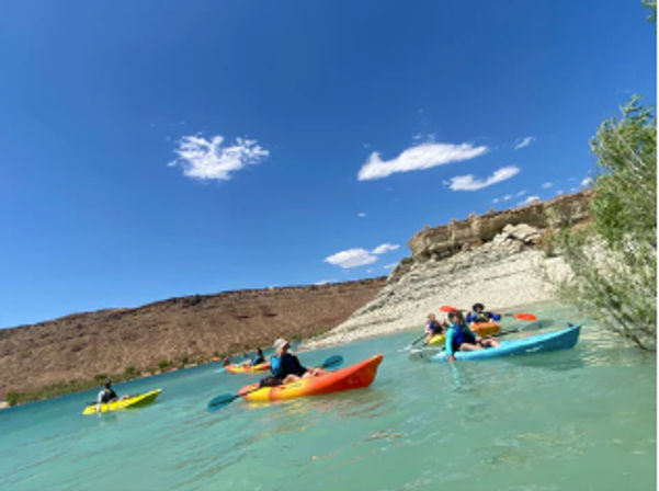 Group of colorful kayaks with paddlers on a sunlit turquoise canyon lake, paddling along sandstone cliffs beneath a clear blue sky – outdoor kayaking adventure.