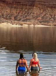 Two women waist-deep in a calm desert canyon lake holding hands, facing red‑rock cliffs and sandy shoreline with mirrored water reflections — outdoor lake scene