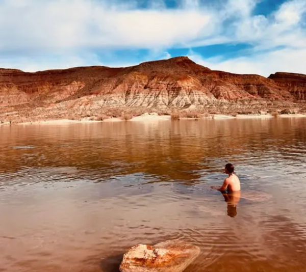 Person wading in a calm brown lake below red sandstone canyon cliffs, with blue sky and the cliffs reflected in the water.