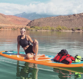 Smiling woman on an orange stand-up paddleboard on a calm desert lake with red-rock hills and distant mountains, a packed backpack and gear beside her.