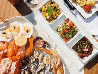 Sunlit seafood platter on ice with whole lobster, crab legs, mussels, oysters, scallops and prawns, a lemon wedge and three small square bowls of colorful salsas and marinated seafood on a wooden table.