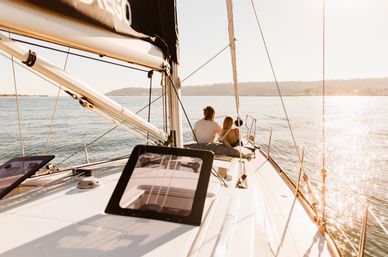 Two people relaxing on a sailboat deck sailing across a sunlit coastal bay at golden hour