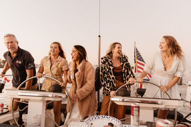 Friends laughing and steering a sailboat at sunset, wearing summer dresses and cozy jackets, holding drinks with an American flag at the stern in a warm golden-hour coastal scene.