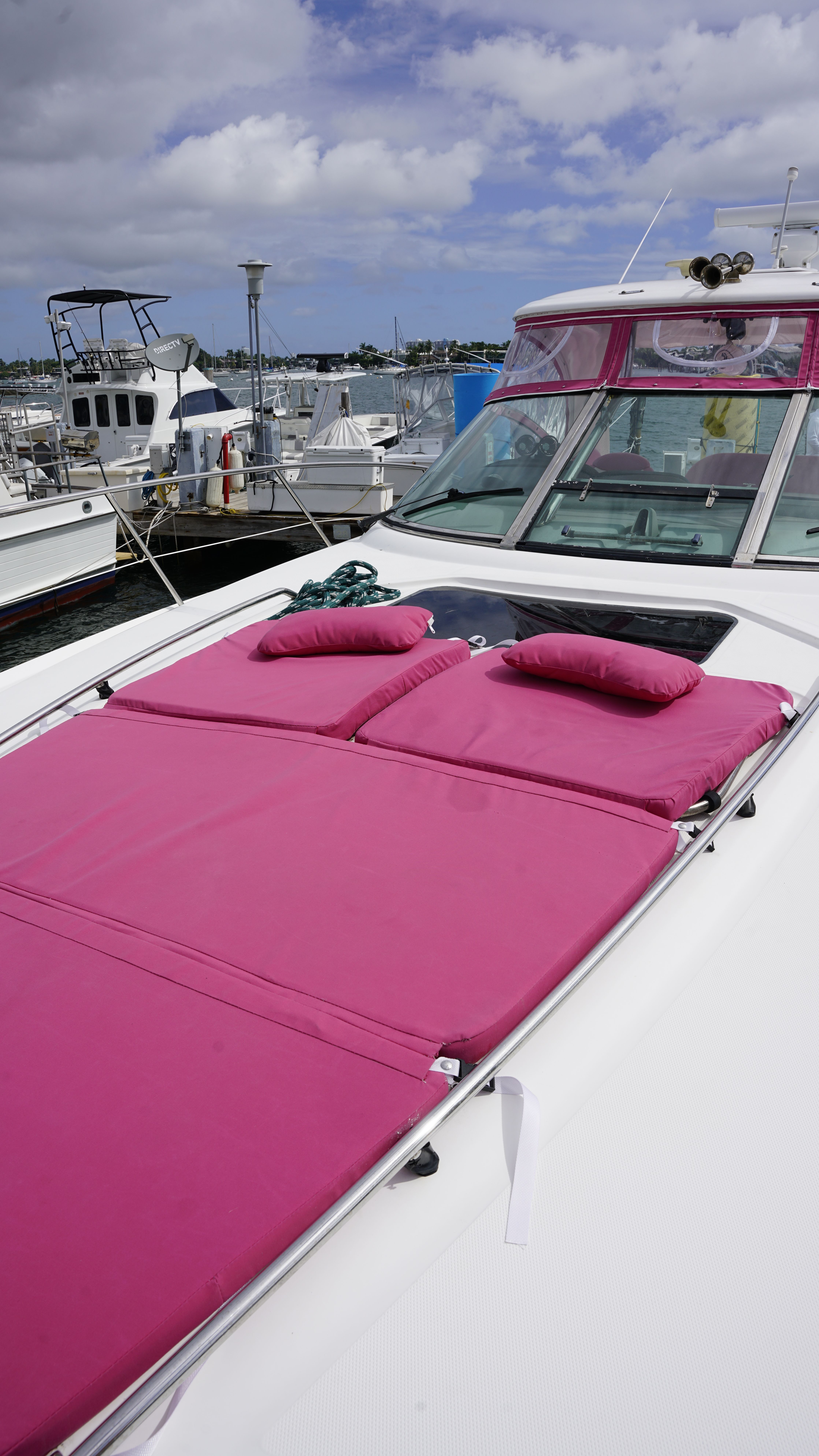 White yacht bow docked at a marina with bright magenta sun pads and pillows on deck, other boats in the harbor and a partly cloudy blue sky.