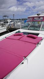 White yacht bow docked at a marina with bright magenta sun pads and pillows on deck, other boats in the harbor and a partly cloudy blue sky.