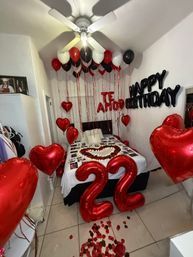 Small bedroom decorated for a romantic 22nd birthday surprise with large red foil '22' balloons, red heart balloons, black 'Happy Birthday' letters on the wall, red/black/white helium balloons at the ceiling, rose petals trail and a heart-shaped arrangement on the bed.