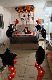 Bedroom decorated for Halloween with orange and black balloons clustered at the ceiling, star-shaped foil balloons, a 'Happy Halloween' wall decal, bed framed by orange petals and LED tea lights, and ghost and jack-o'-lantern pillows.