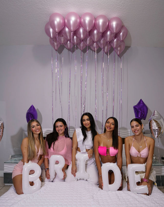Indoor bachelorette party: five women seated on a bed holding white letter balloons that spell BRIDE, wearing pink and white outfits under a cluster of pink helium balloons and purple star balloons.