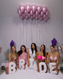Indoor bachelorette party: five women seated on a bed holding white letter balloons that spell BRIDE, wearing pink and white outfits under a cluster of pink helium balloons and purple star balloons.