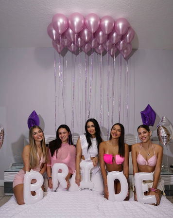 Indoor bachelorette party: five women seated on a bed holding white letter balloons that spell BRIDE, wearing pink and white outfits under a cluster of pink helium balloons and purple star balloons.