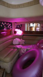 Pink-lit yacht cabin lounge with curved tan leather seating, floral wall panel, neon pink sign, shell and star-shaped pillows, sequin throw and large inflatable pink tube.