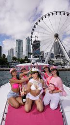 Cheerful bachelorette boat party with the bride-to-be in a white sash and captain hat holding champagne, surrounded by friends in pink sailor hats and swimsuits on a yacht with a waterfront city skyline and a large Ferris wheel in the background.