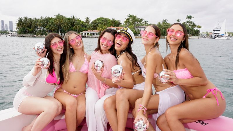Six women in pink swimwear and matching pink sunglasses laughing on a boat at a Miami waterfront, holding mirrored disco-ball drink cups