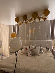 Modern bedroom decorated for a birthday with gold and white helium balloons floating to the ceiling, long metallic ribbons holding small photo cards above a neatly made bed and a 'Happy Birthday' wall banner, plus a large white balloon with a Spanish congratulatory message.
