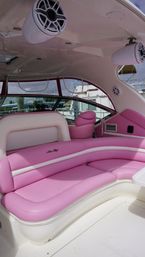 Curved pink-and-white yacht interior with plush banquette seating, pink captain’s chair, overhead marine speakers and marina visible through wraparound windows