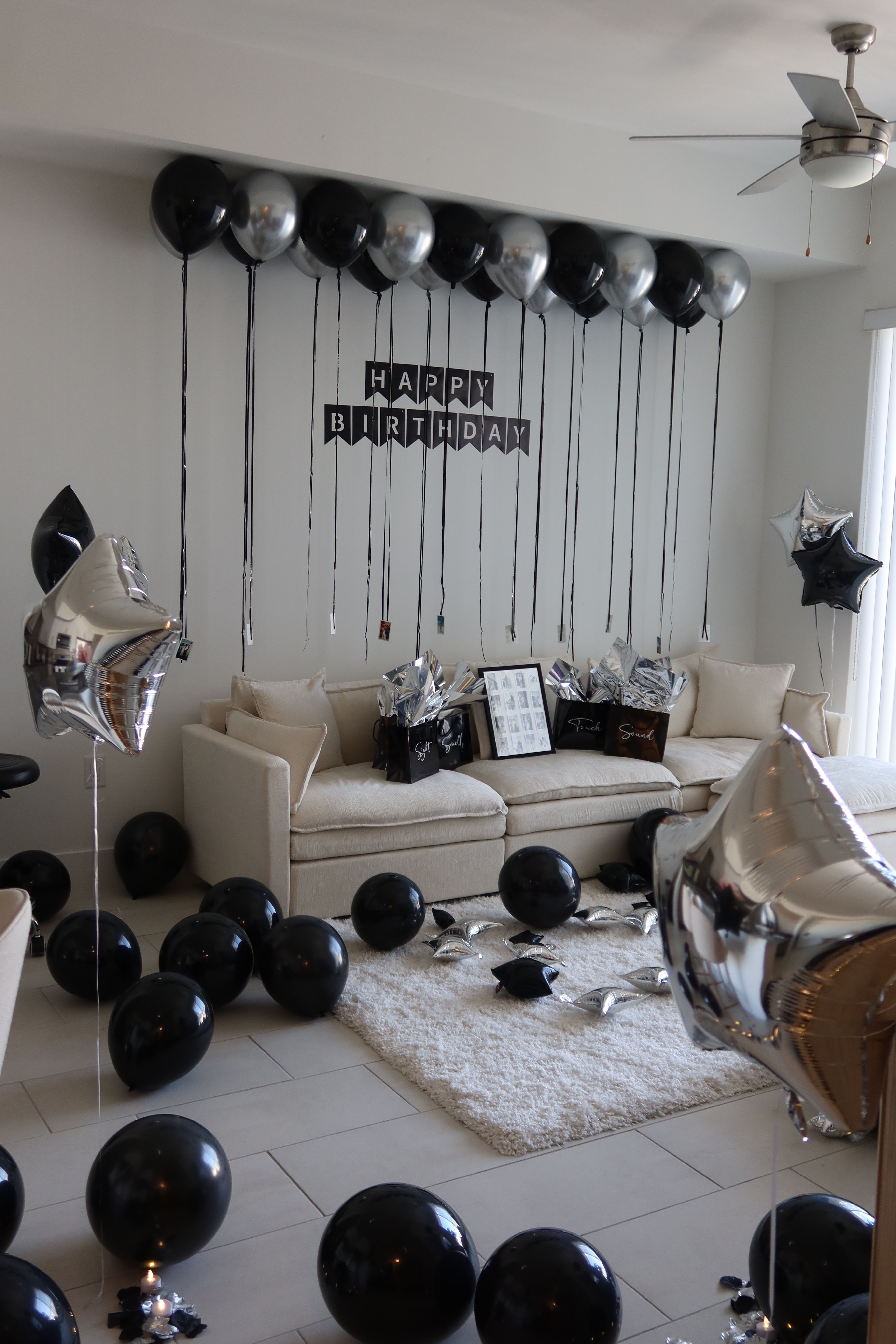 Modern living room decorated for a black-and-silver birthday: beige sofa with gift bags, a 'Happy Birthday' banner, ceiling-tethered black and metallic silver balloons and star-shaped mylar balloons scattered on the floor and rug.