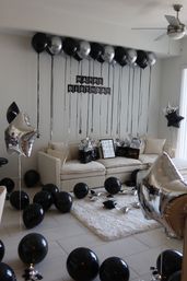 Modern living room decorated for a black-and-silver birthday: beige sofa with gift bags, a 'Happy Birthday' banner, ceiling-tethered black and metallic silver balloons and star-shaped mylar balloons scattered on the floor and rug.