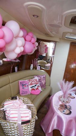 Pink-themed party setup inside a yacht cabin with pink-and-white balloon garland, 'Cherry Bomb' pillows, gift basket, and satin-covered table with photo props — playful celebration decor