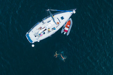 Aerial drone shot of a white sailboat anchored in deep blue water with a red dinghy alongside and two swimmers splashing nearby, summer boating scene