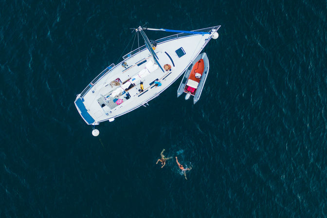 Aerial drone shot of a white sailboat anchored in deep blue water with a red dinghy alongside and two swimmers splashing nearby, summer boating scene