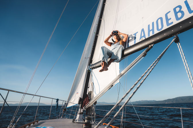 Person in light overalls perched on a sailboat boom using binoculars, sailing across deep-blue ocean with distant coastline under a clear sky.