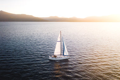 Aerial view of a white sailboat with blue-trimmed sails gliding across calm open water toward a mountain-lined horizon bathed in warm sunset light